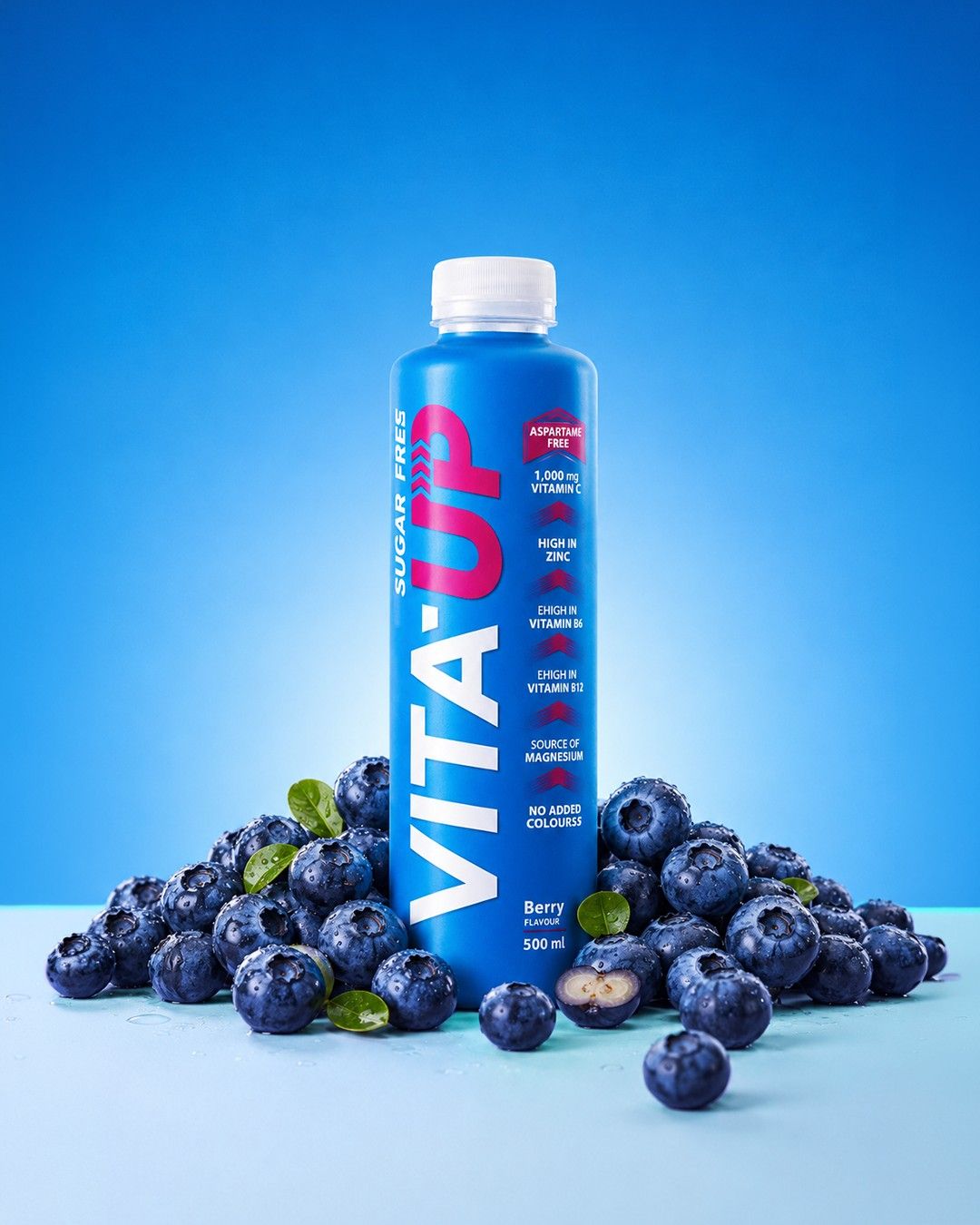 A Berry bottle of Vita-Up standing in a pile of fresh blueberries on a blue backdrop