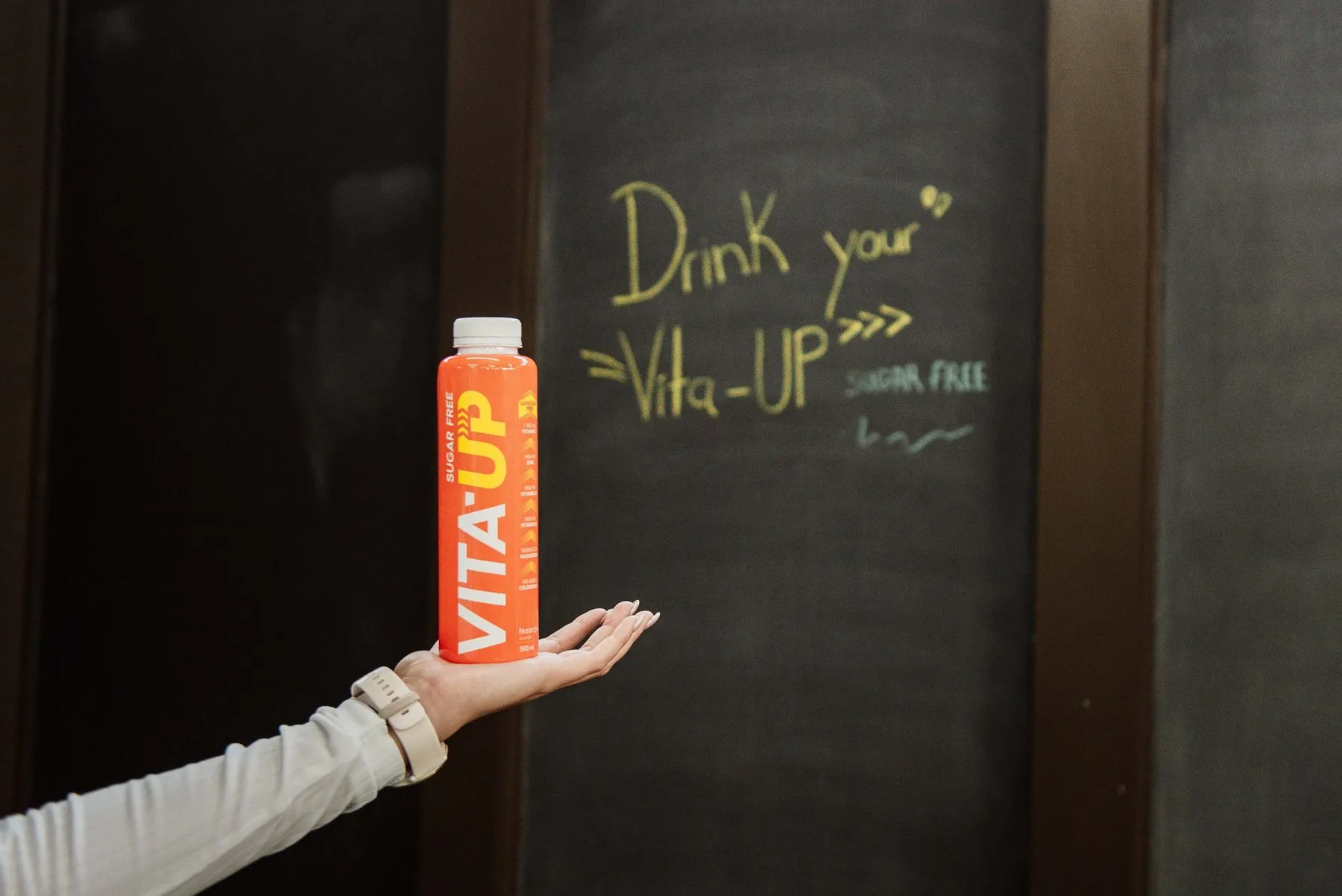 Hand balancing a Naartjie bottle in front of a chalkboard that says Drink your Vita-Up, sugar free